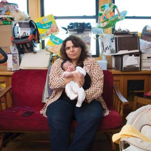 A woman with brown curly hair and a cardigan is sitting on a couch and holding her baby, who has dark hair and wears a white longsleeve footie onesie. Piles of boxes, baby supplies, and furniture surround her on all sides.