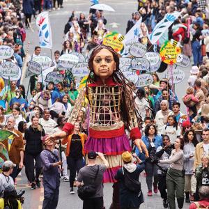 An 11-foot puppet designed to look like a Syrian child is surrounded by a crowd with signs advocating for relief for refugees.