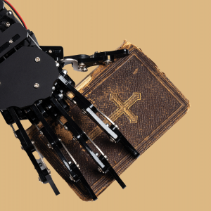 The picture shows a robotic hand holding a Bible on a tan/gold background 