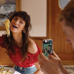 A woman is pictured holding up a fork full of noodles to her smiling mouth while posing for a photo