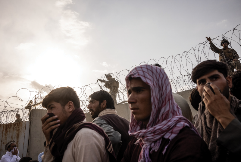 Afghan men stand before a concrete wall topped with barbed wire and a U.S. soldier