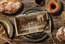 The image shows a table from a bird's-eye-view, with a loaf of bread, a pitcher, and a fish. In the middle there is a platter with an image of the Last Supper, being cut apart by a fork. 