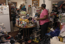 A woman stands in a kitchen with objects piled all over a table and along the walls 