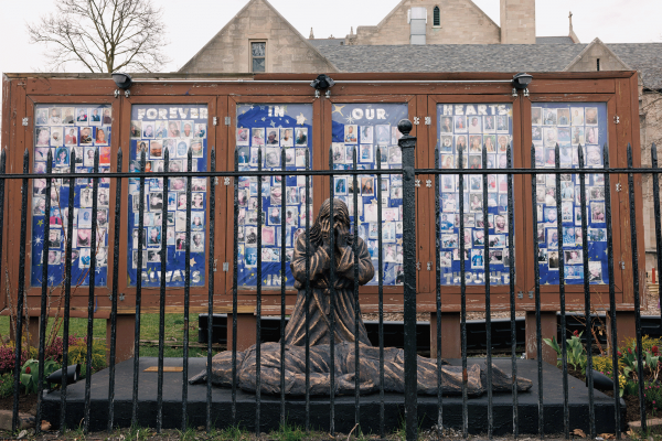 The image shows a sculpture of a weeping person kneeling over a dead body with a display in the back showing faces of people killed by gun violence. 