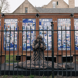 The image shows a sculpture of a weeping person kneeling over a dead body with a display in the back showing faces of people killed by gun violence. 