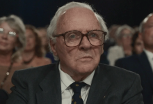 The picture shows an old white man with glasses wearing a suit looking at something, semi-puzzled or surprised. There are people behind him. 