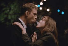 A white man wearing a suit blows smoke into the mouth of a young white woman